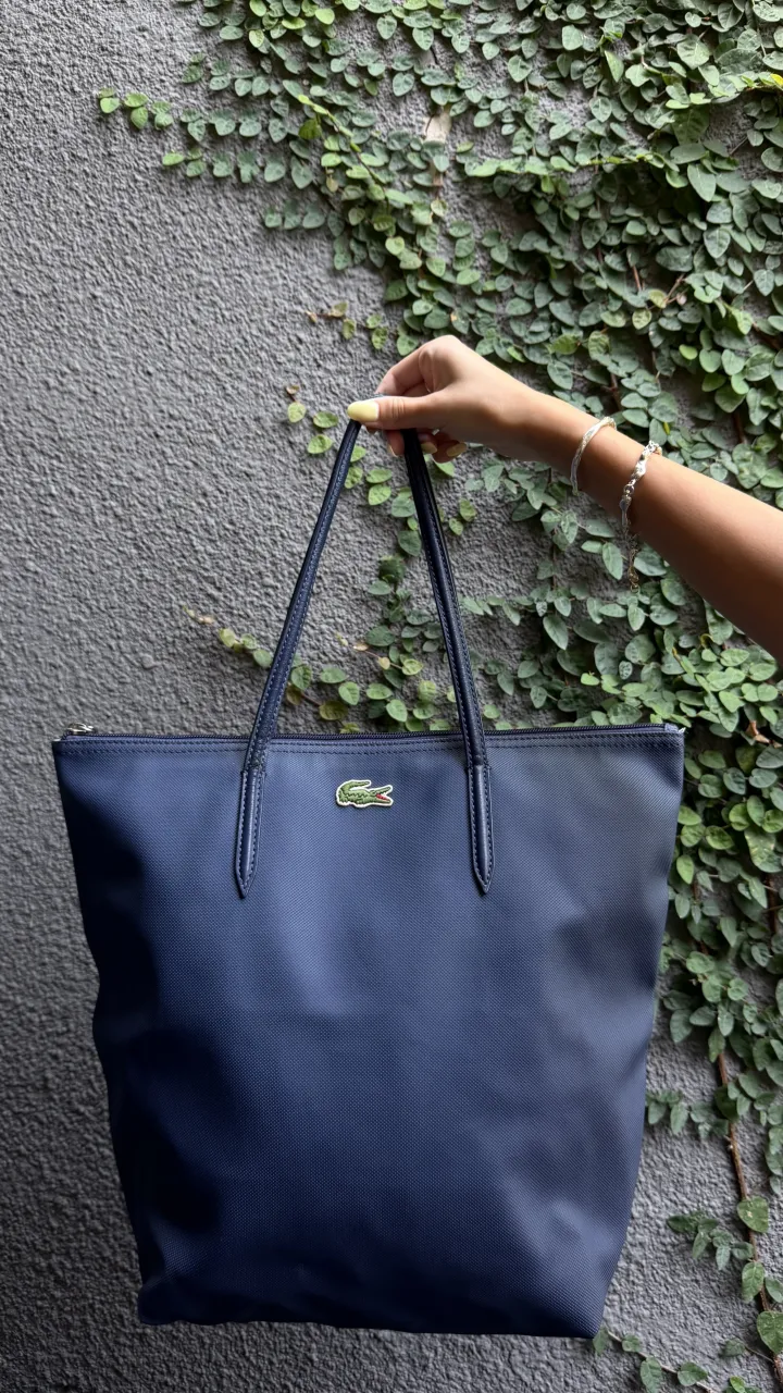 Bolso tote azul marino con logo de cocodrilo verde bordado en el frente. Tiene dos asas y cierre superior.