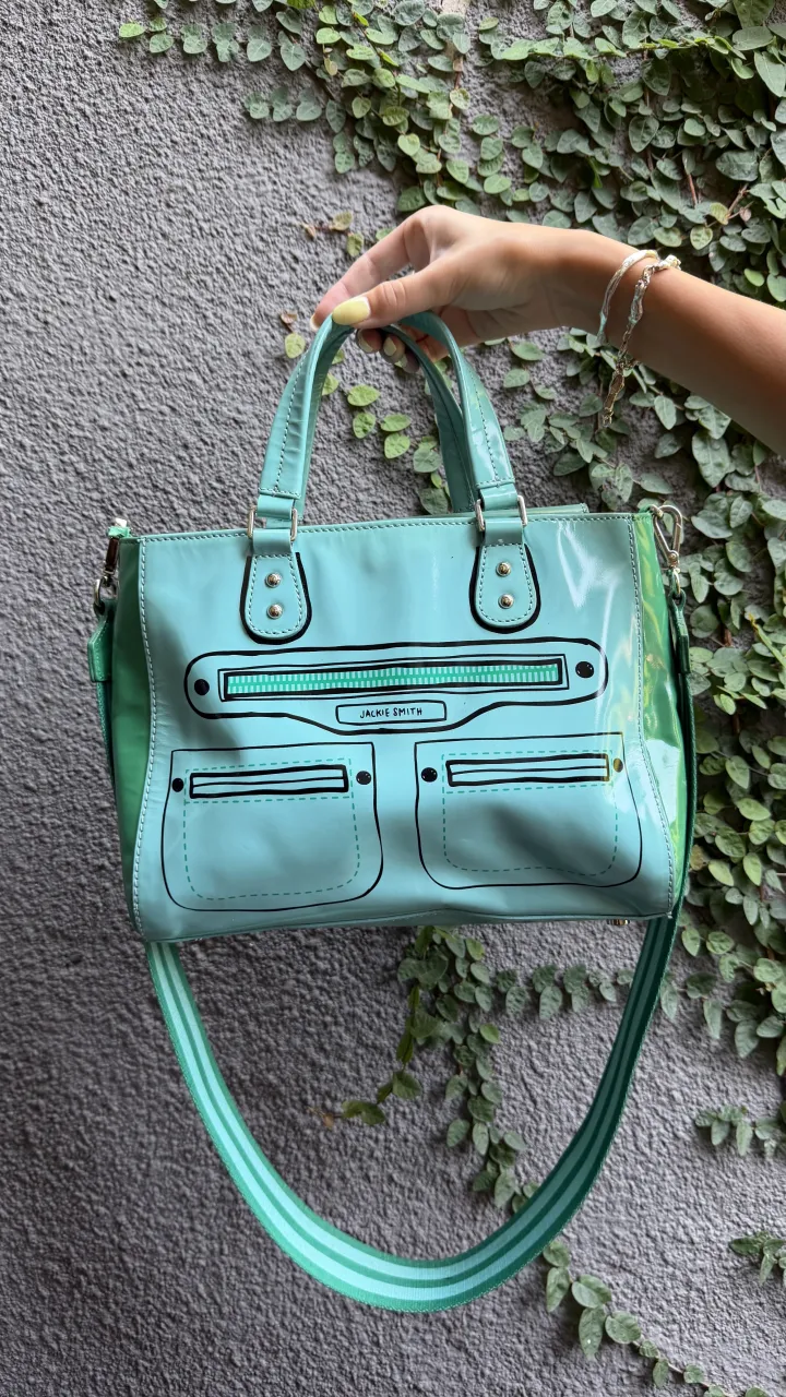 Bolso tote color celeste con detalles de diseño que simulan bolsillos y cierre. Posee dos manijas cortas y una correa larga desmontable con detalles de rayas en dos tonos de celeste. En el frente tiene un recuadro con el nombre 'JACKIE SMITH'.