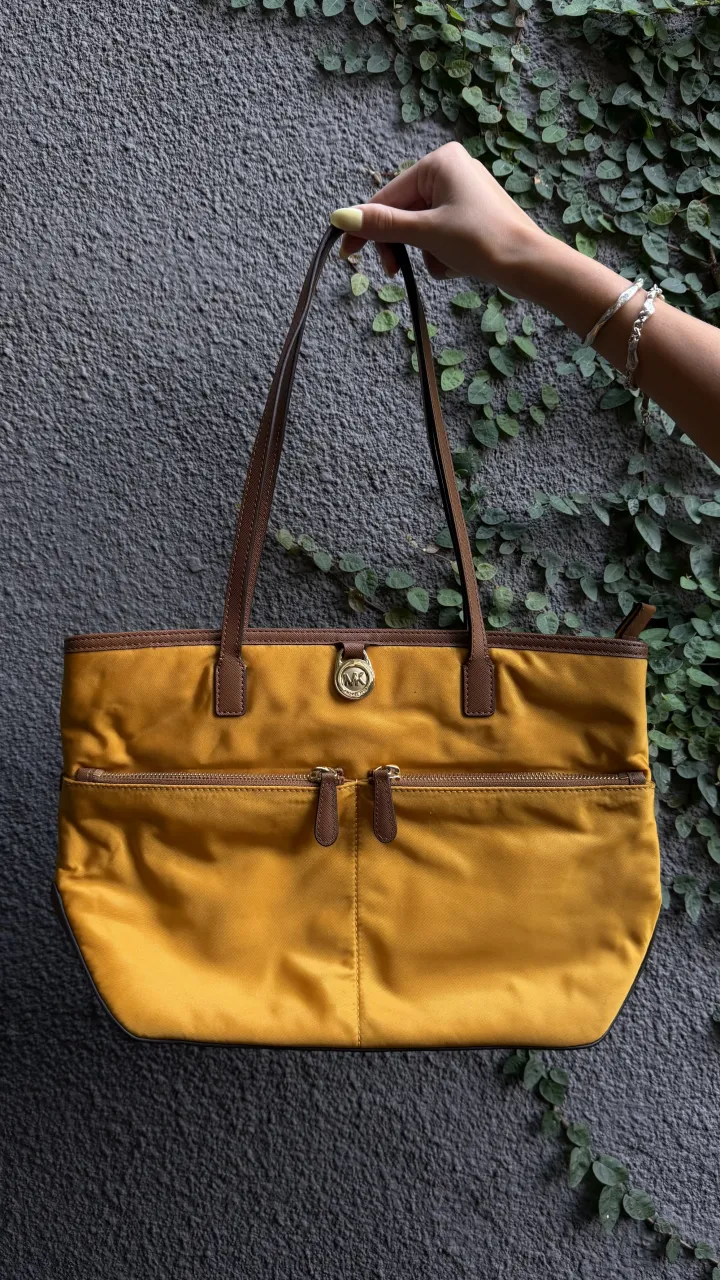 Bolso tote amarillo con dos bolsillos frontales con cierre y tiradores de cuero marrón. Las asas son de cuero marrón y tiene un logo metálico dorado en el frente.