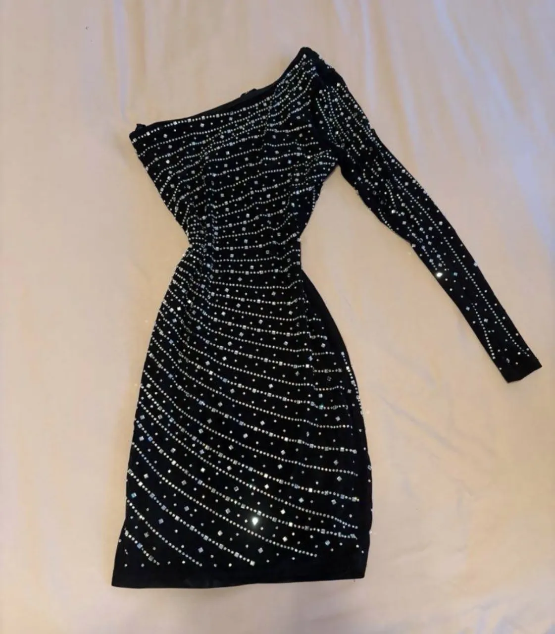 Vestido negro con un solo hombro, manga larga y detalles de pedrería brillante en toda la prenda. Posee fruncido en un lateral de la falda.