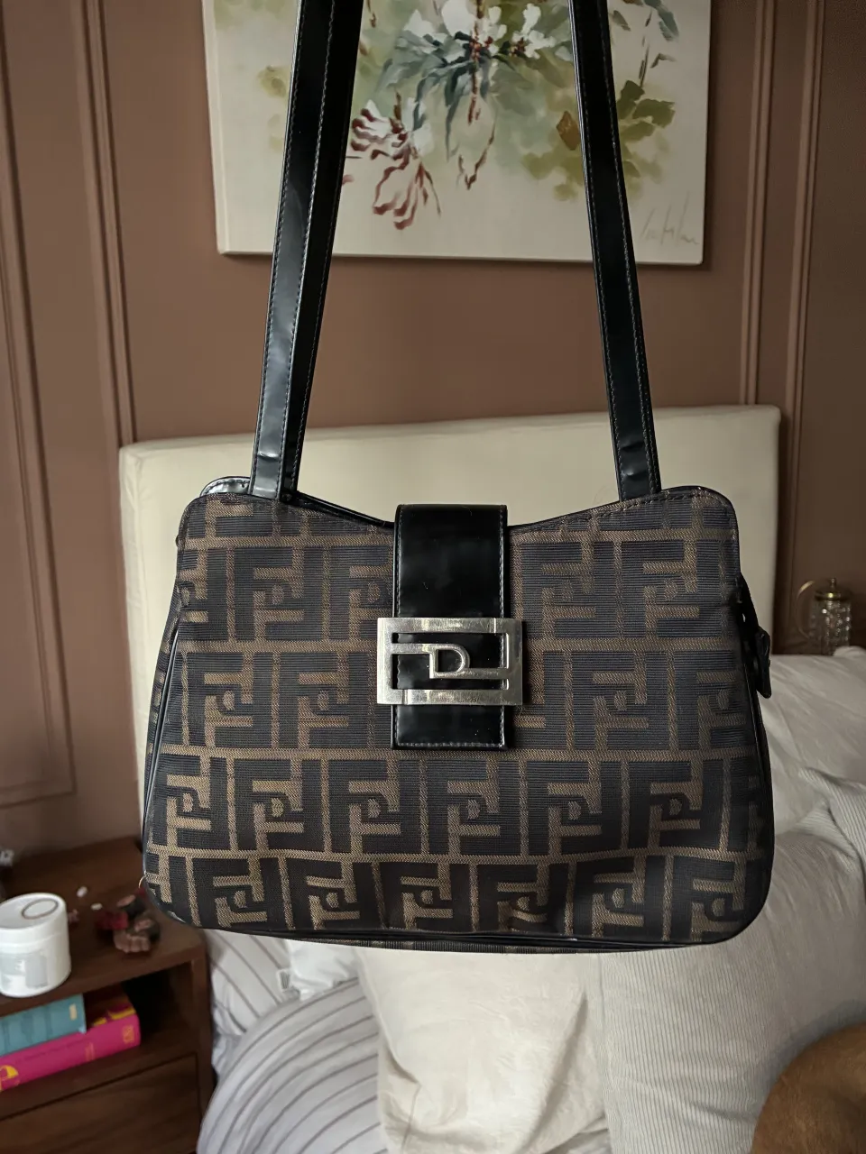 Bolso de hombro con estampado monograma en tonos marrón y negro. Posee un bolsillo exterior con cierre y un detalle metálico plateado con logo en el frente.