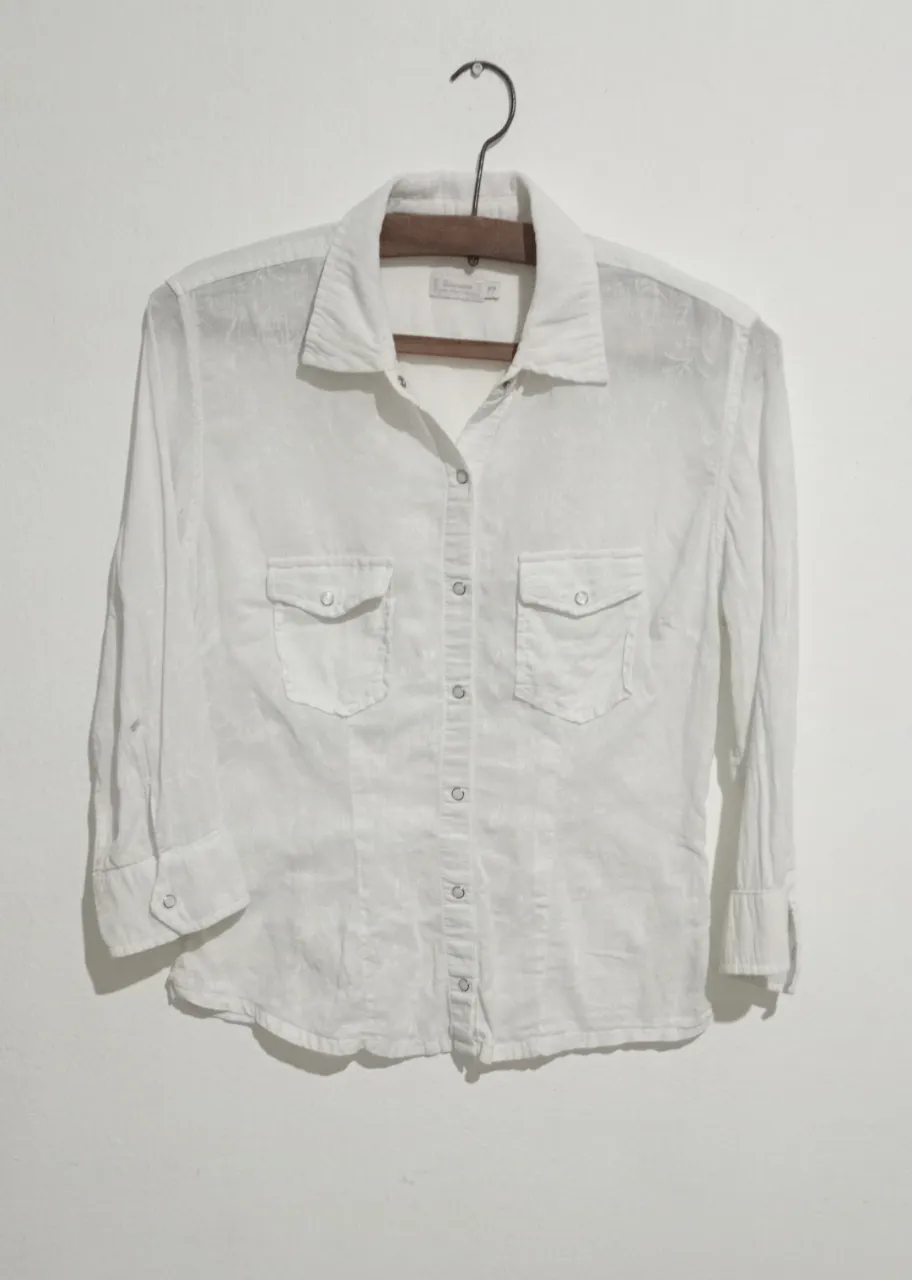 Camisa blanca de manga larga con botones al frente y dos bolsillos en el pecho. Posee un estampado floral sutil. La etiqueta indica la marca 'Wanama'. Talle S. Botones a presión.
