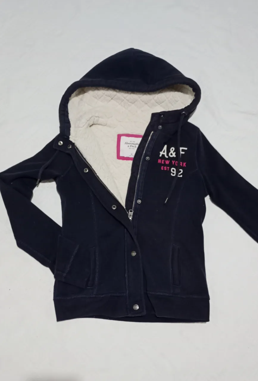 Campera negra con capucha, forrada en sherpa blanco (no tiene forro en la parte interna de los brazos). Posee cierre y broches metálicos en el frente. Estampa de la marca en el frente. Marca Abercrombie&Fitch. Elastizada. Talle XS. DETALLES DE USO: Mancha en el puño de manga (ver última foto). Medidas aprox. en plano: hombros: 37 cm, sisa 43 cm, largo 55 cm.