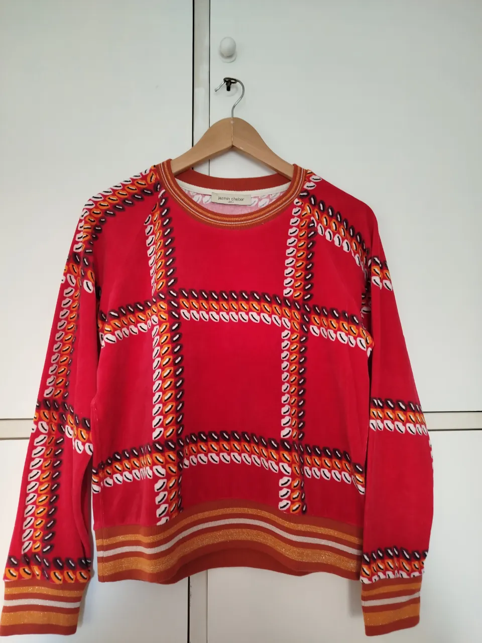 Sweater Jazmín Chebar de mujer con diseño geométrico en tonos rojos, naranjas y blancos. Cuello redondo y puños con detalles dorados. Ideal para un look casual y moderno.