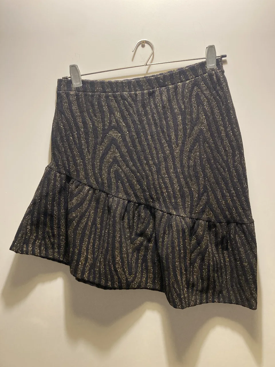 Falda irregular de mujer de Jazmin Chebar, con estampado de cebra en tonos negro y dorado. Tiene un detalle de volado en el borde inferior y un botón dorado con el logo de la marca. Ideal para un look elegante y sofisticado. La tela es elastizada. Talle M/L.