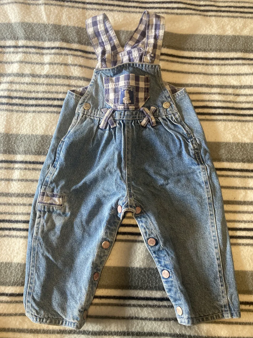 Mono corto de jean para bebé, con detalles a cuadros en la pechera y tirantes. Diseño con botones en la entrepierna para facilitar el cambio de pañales. Marca Coniglio. 6 meses
