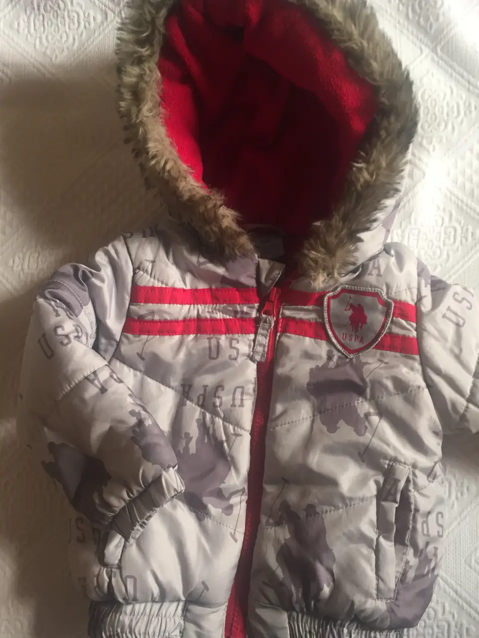 Campera de abrigo para bebé, con capucha y forro polar rojo. Diseño camuflado en tonos grises y detalles en rojo. Con el logo de U.S. Polo Assn. en el pecho. Ideal para mantener a tu bebé abrigado y a la moda.