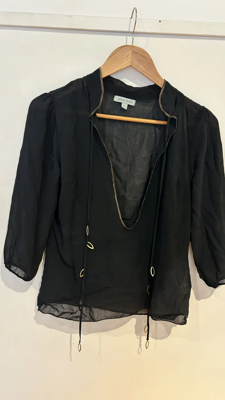 Blusa negra transparente con detalles dorados en el cuello y cordones para atar. Mangas 3/4 con un corte sutil y elegante. Perfecta para un look sofisticado.