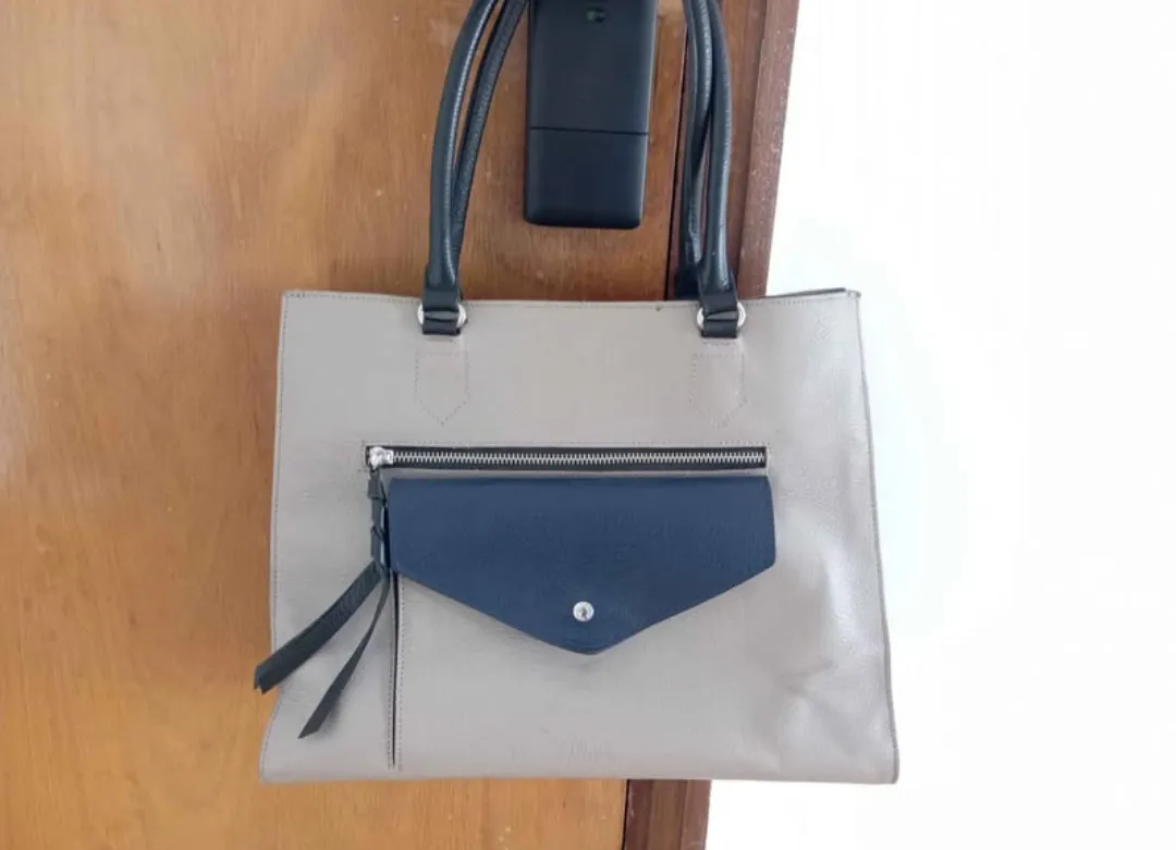 Cartera tote bag de cuero sintético en color gris con detalles en azul marino. Posee un bolsillo frontal con solapa y cierre. Ideal para el trabajo o uso diario.