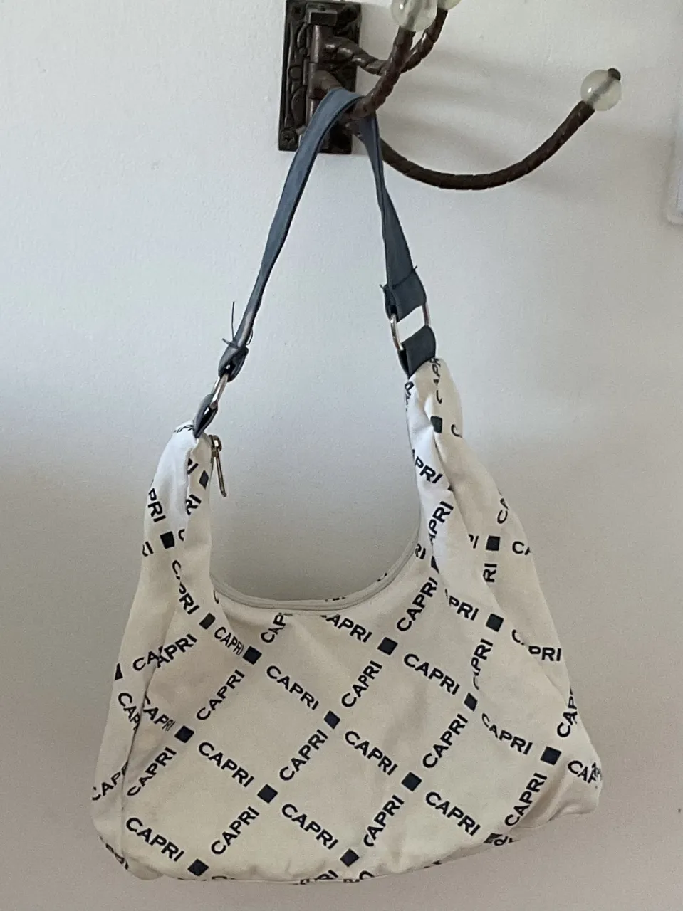 Bolso blanco con estampa de la palabra "CAPRI" en negro, formando un patrón de cuadros. Tiene una correa de hombro ajustable en color gris oscuro. Ideal para el día a día.
