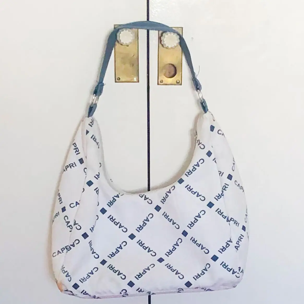 Cartera tipo baguette blanca con estampado repetido de la palabra "CAPRI" en azul. Ideal para llevar al hombro, con un diseño moderno y práctico.