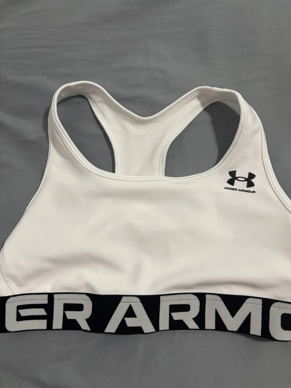 Top deportivo blanco de la marca Under Armour, ideal para entrenamientos. Posee el logo de la marca en el frente y una banda elástica con el nombre de la marca en la cintura.