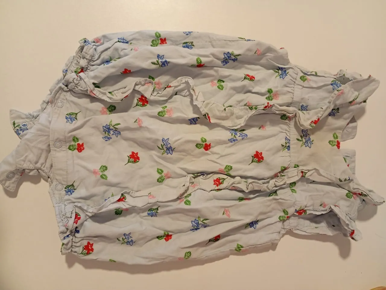 Mono de bebé estampado con flores rojas, azules y rosas sobre fondo celeste. Posee volados en los hombros y broches en la parte inferior para facilitar el cambio.