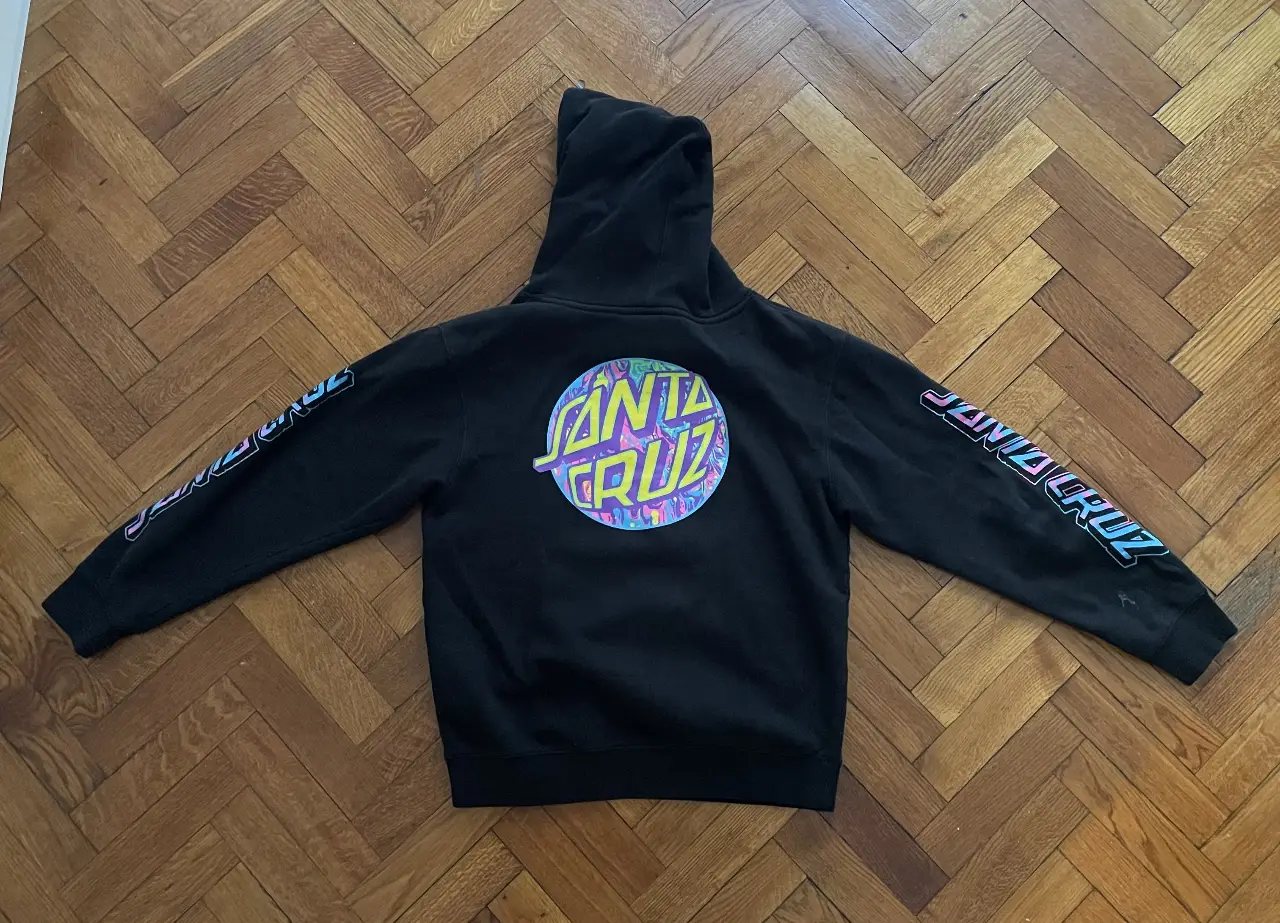 Buzo canguro negro de la marca Santa Cruz, con capucha y bolsillo frontal. Estampado del logo de la marca en el frente y en las mangas. Ideal para un look urbano y casual.