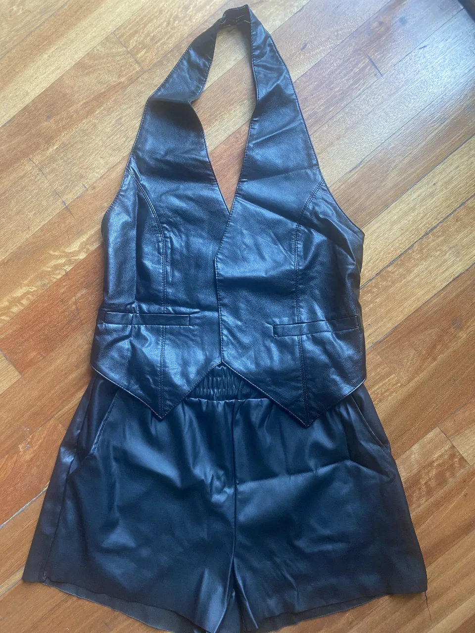 Conjunto de short y top estilo chaleco de cuero sintético negro. El top es tipo halter con escote en V pronunciado y detalles de bolsillos falsos. El short tiene cintura elastizada y corte suelto. Ideal para un look audaz y moderno.