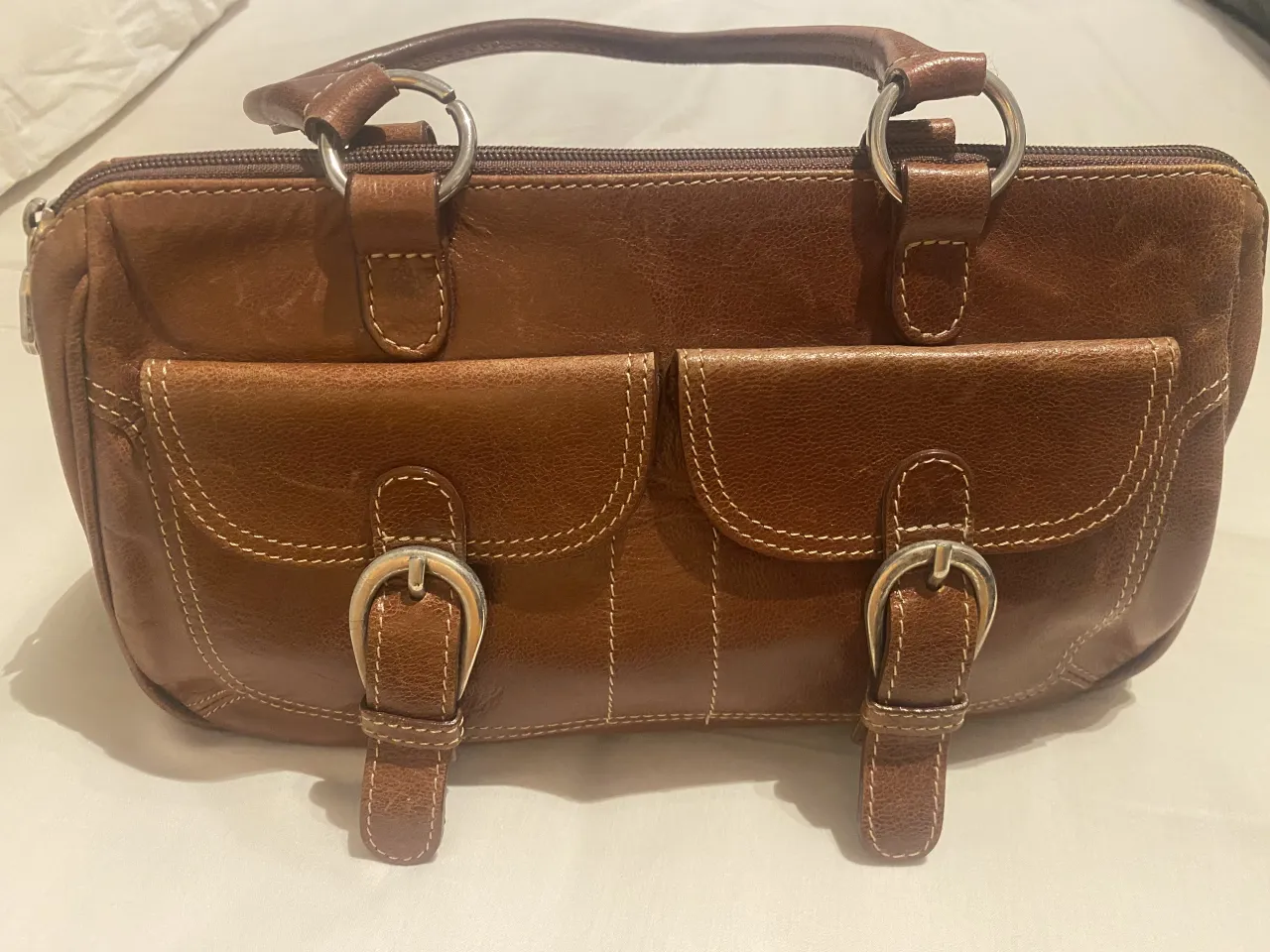 Cartera marrón de cuero con dos bolsillos frontales con hebillas plateadas. Posee dos manijas superiores y un cierre superior. Medidas 32 x 17.  Ideal para uso diario.