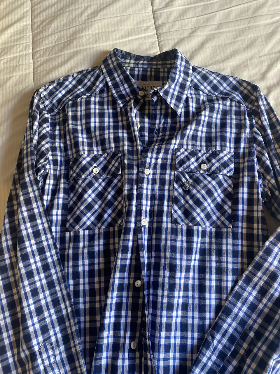 Camisa a cuadros azul y blanca de manga larga, con dos bolsillos delanteros y botones blancos. Ideal para un look casual y versátil. Talle 42/L