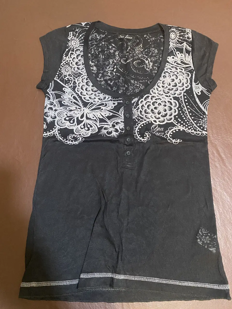 Remera negra con estampado floral en blanco en la parte delantera y trasera. Cuello con botones y detalle de encaje en la espalda. Ideal para un look casual y moderno.