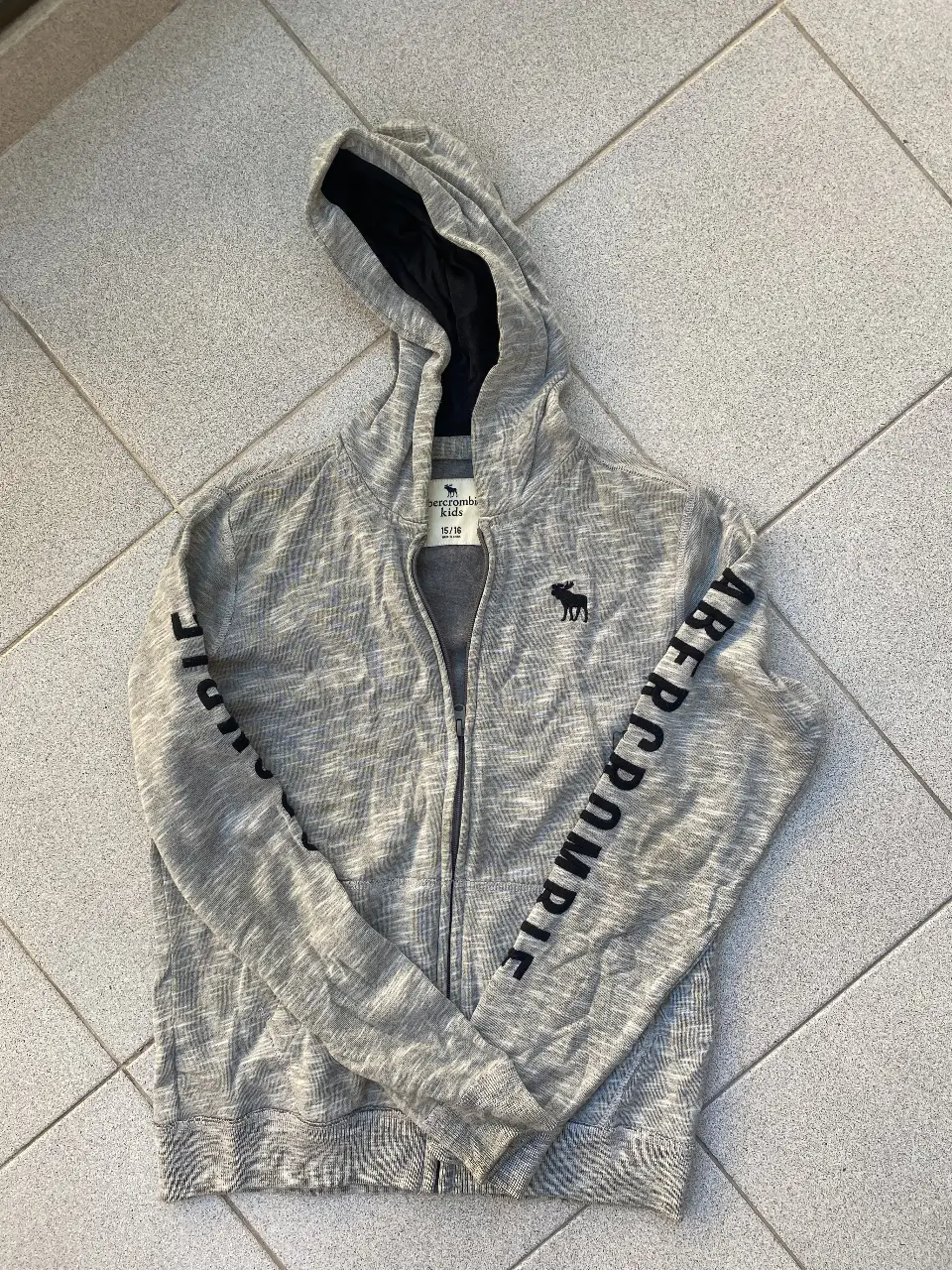 Buzo con cierre y capucha para niño, de la marca Abercrombie Kids. Con mangas estampadas con el nombre de la marca y logo en el pecho. Talle 10/14