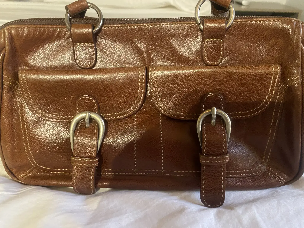 Cartera marrón de cuero con dos bolsillos frontales con hebillas plateadas. Posee dos manijas superiores y un cierre superior. Ideal para uso diario.
