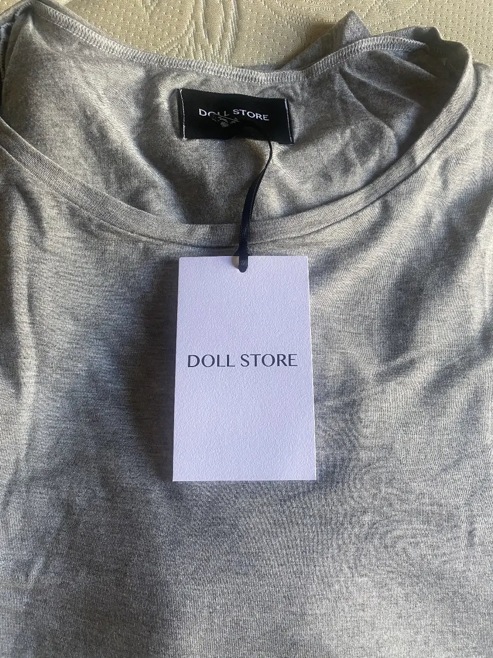 Remera gris de manga corta, con cuello redondo y etiqueta de la marca 'DOLL STORE'. Ideal para un look casual y cómodo y amplio. SIN USO CON ETIQUETA 