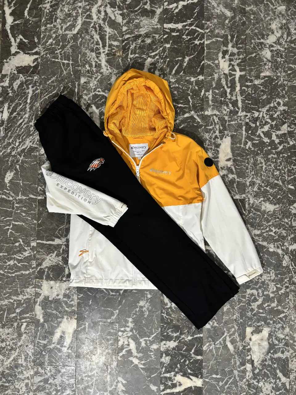 Conjunto deportivo Discovery Expedition para hombre. Incluye campera rompevientos con capucha en amarillo y blanco, y pantalón de jogging negro con detalles de la marca. Ideal para actividades al aire libre y un look casual.