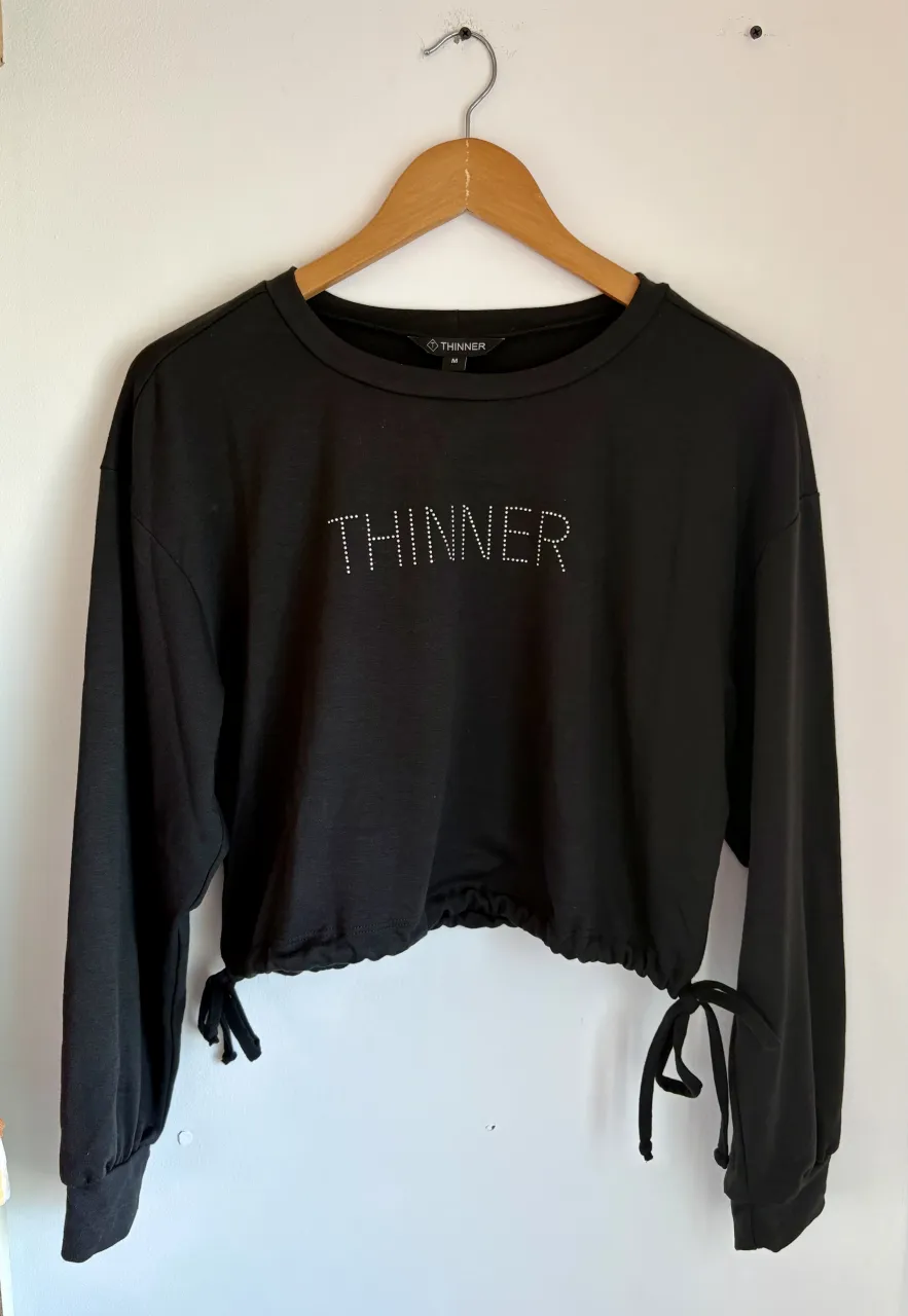Remera negra manga larga, crop, con cordones ajustables en el ruedo y la palabra "THINNER" estampada con strass en el frente.