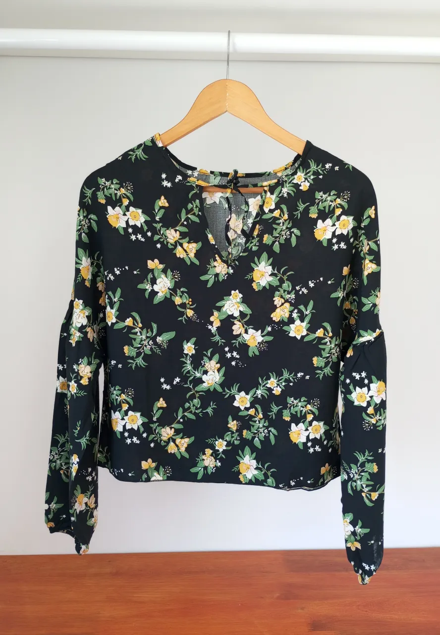 Blusa estampada con flores en tonos amarillos y blancos sobre fondo negro. Posee mangas largas con un detalle de volado en el puño y un escote en V con amarre. Ideal para un look casual y femenino.
