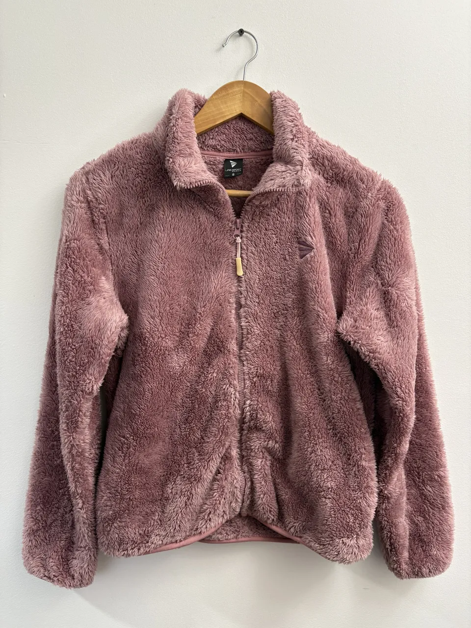 Campera de plush color rosa con cierre completo y cuello alto. Tiene un logo pequeño bordado en el frente.