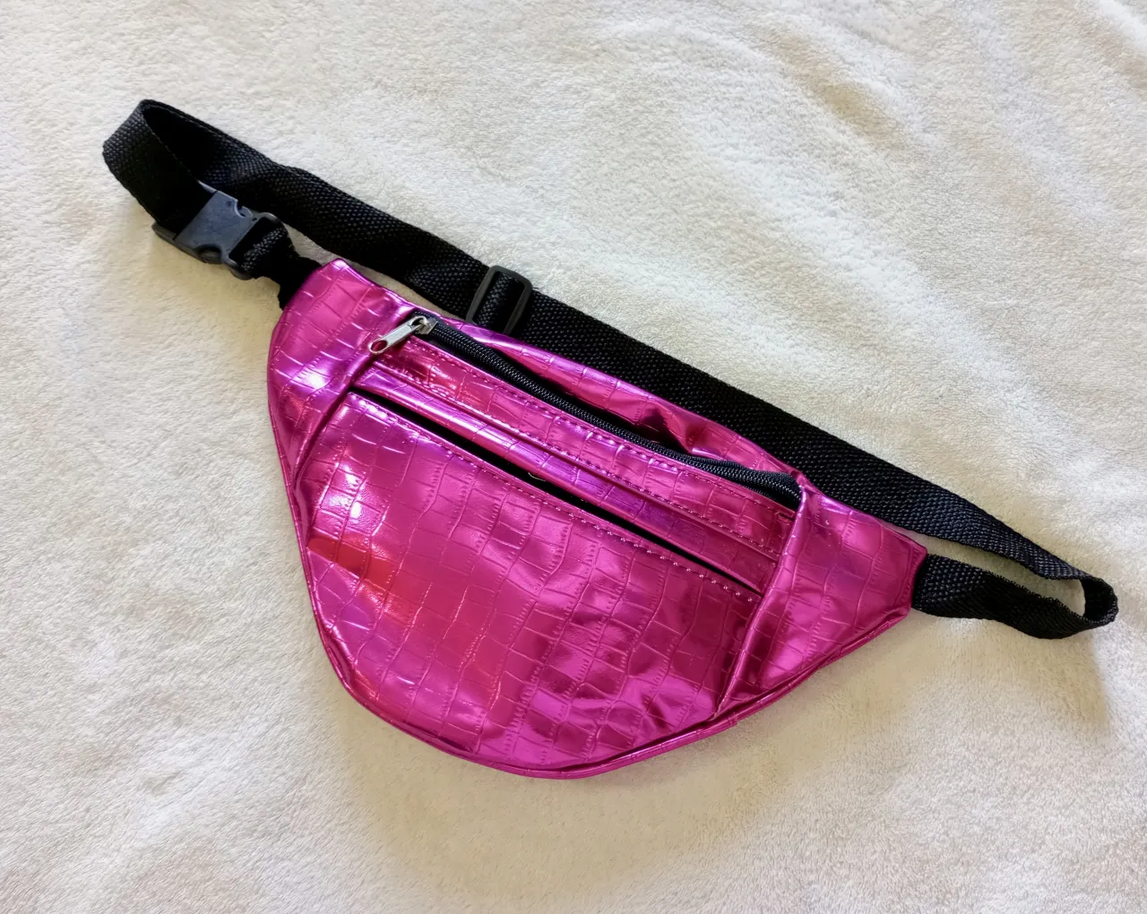 Riñonera fucsia metalizada con textura de cocodrilo. Tiene dos compartimentos con cierre y correa ajustable negra.