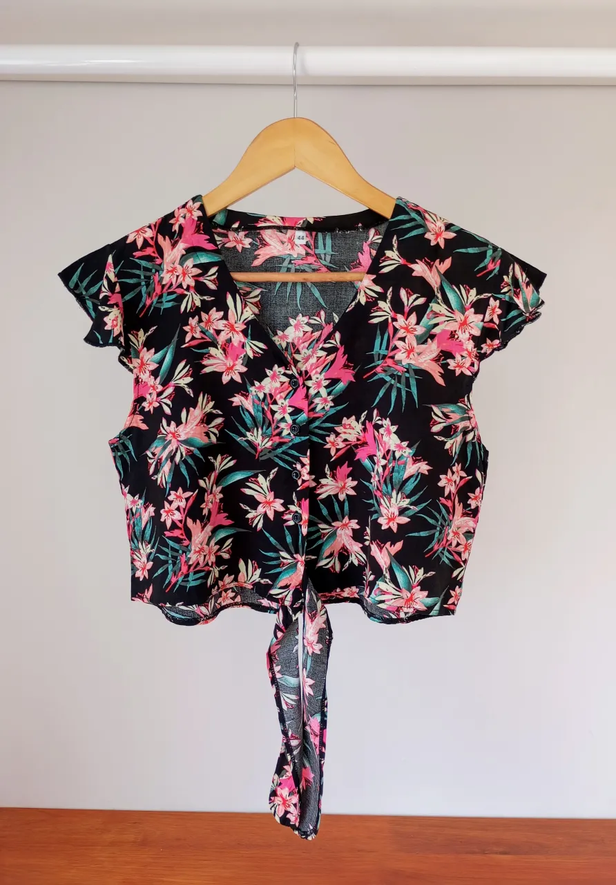 Camisa crop top estampada con flores y hojas en tonos rosas, verdes y blancos sobre fondo negro. Posee cuello en V, botones al frente y lazo para atar en la cintura.