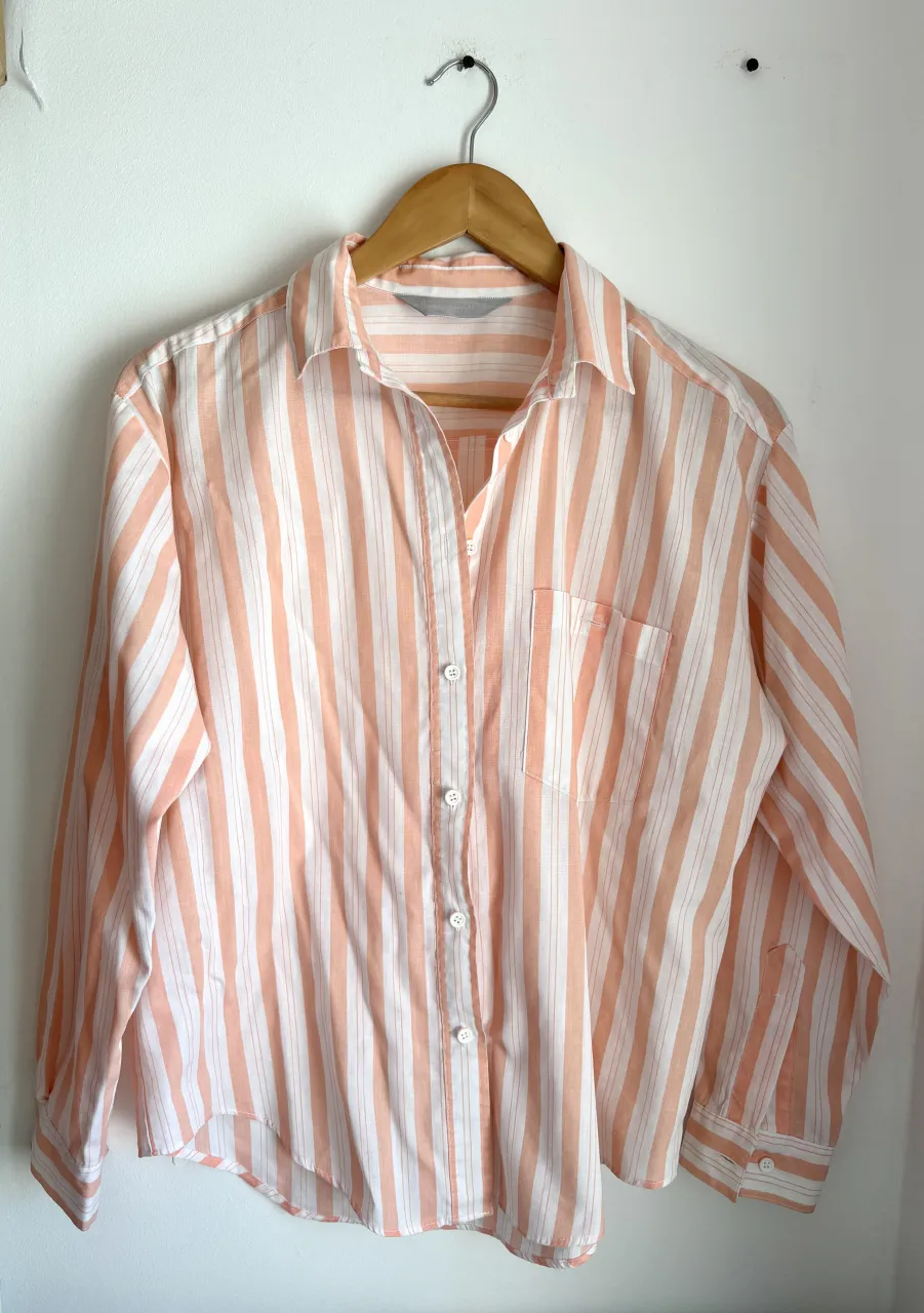 Camisa a rayas verticales en tonos blanco y salmón. Posee cuello camisero, manga larga y botones al frente. Tiene un bolsillo en el pecho del lado izquierdo.