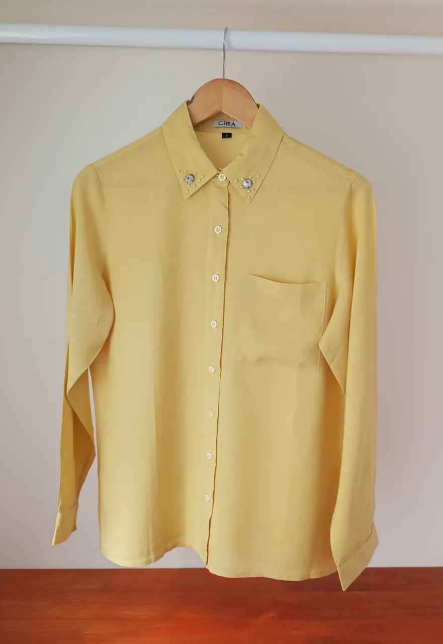 Camisa de manga larga color amarillo. Cuello con detalles de pedrería y perlas. Posee un bolsillo en el frente.