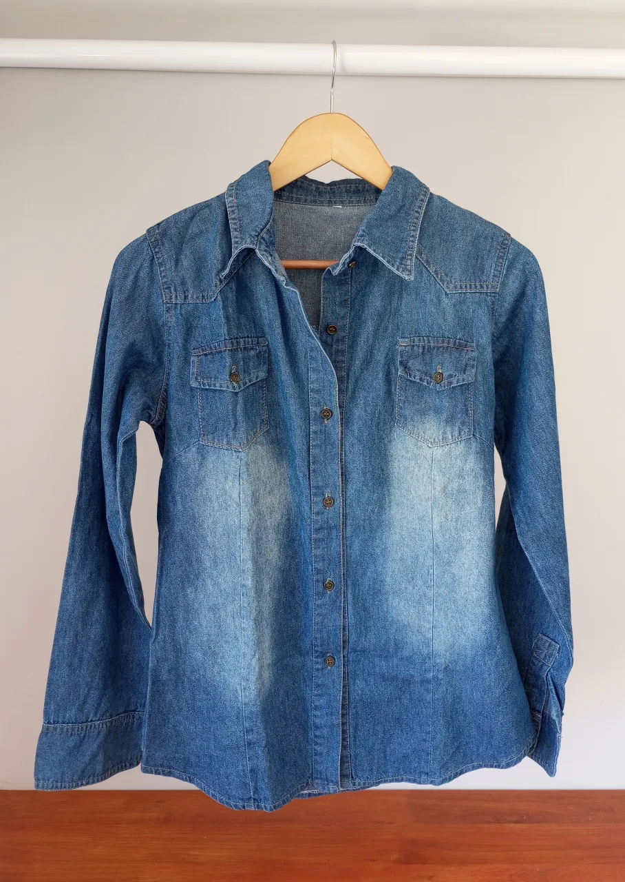 Camisa de jean azul con lavados desgastados. Posee cuello camisero, botones al frente y dos bolsillos con solapa en el pecho. Mangas largas con puño.
