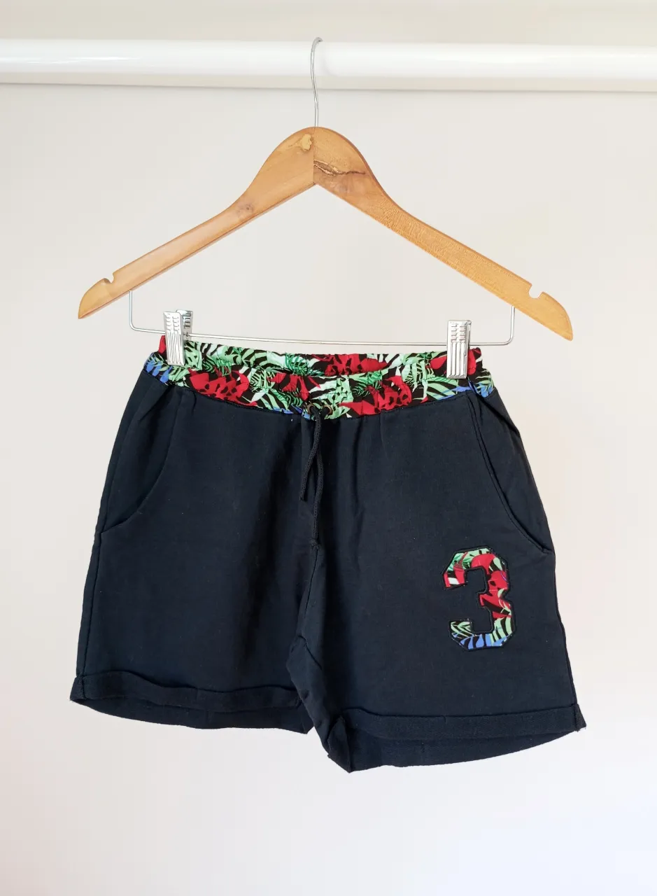 Shorts negros con cintura elastizada y cordón. Presentan un estampado floral en la cintura y un aplique de número '3' con el mismo estampado en la parte inferior izquierda.