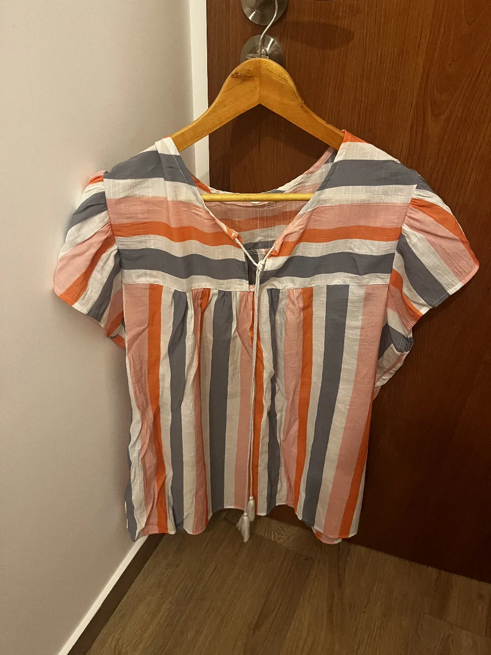 Remera de rayas verticales en tonos coral, gris y blanco. Posee escote en V con detalle de cordón y borla. Mangas cortas con un leve fruncido en los hombros.