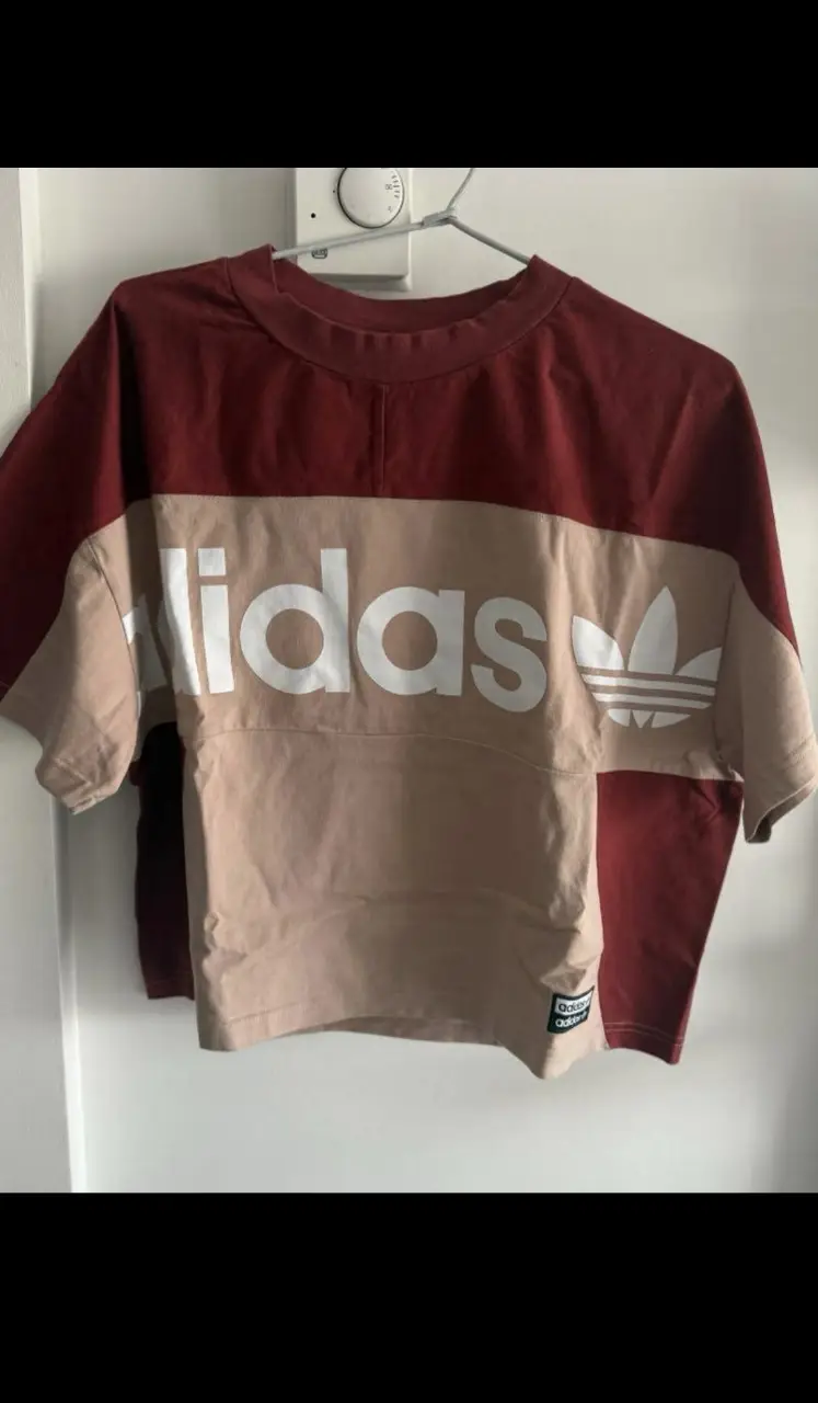 Remera Adidas de manga corta para mujer, con diseño en bloques de color rojo y crudo. Presenta el logo de la marca en blanco en el frente. Ideal para un look deportivo y casual. Talle S/M