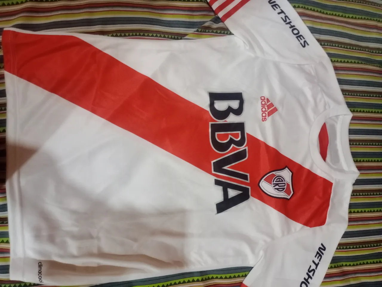 Camiseta de fútbol blanca con banda roja diagonal en el frente y espalda. Logo de Adidas y escudo del club en el frente. Inscripción 'BBVA' en el frente y 'BBVA Francés' en la espalda. Inscripción 'NETSHOES' en las mangas.