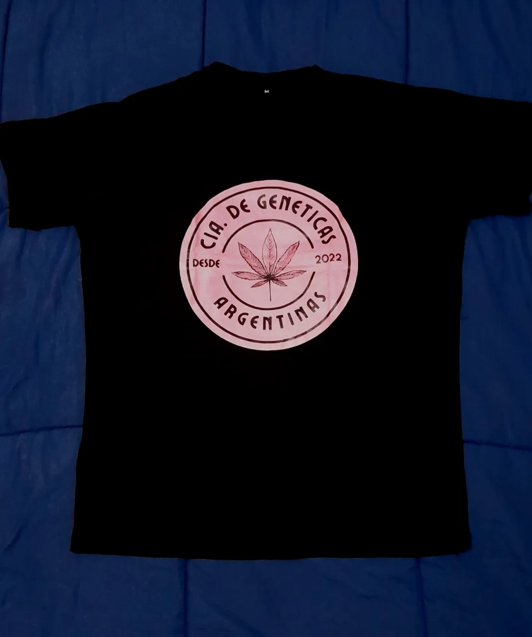 Remera negra con estampa circular en color rosa viejo. La estampa tiene una hoja de maría en el centro y el texto "CIA. DE GENETICAS ARGENTINAS DESDE 2022".

66cm de largo
55cm de axila a axila
