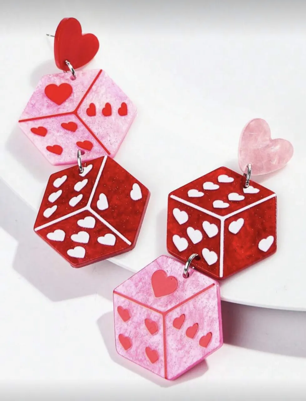 Aritos colgantes con forma de dado y corazones. Los dados son de color rojo y rosa con detalles de corazones blancos y rojos. Tienen un corazón rojo en la parte superior.