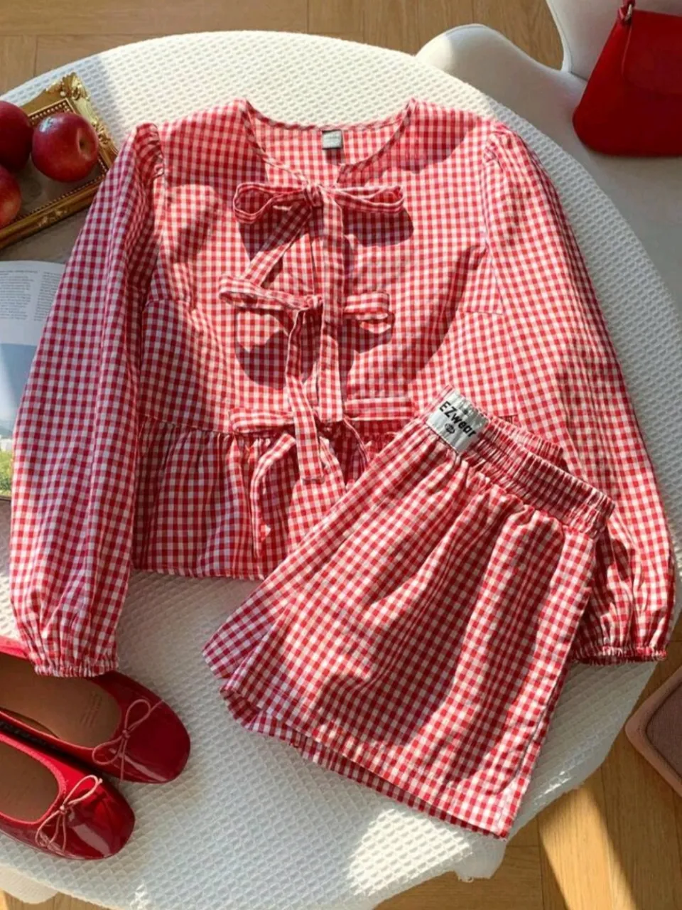 Conjunto de dos piezas a cuadros escoceses en rojo y blanco. La blusa tiene mangas largas abullonadas, cuello redondo con lazo y volado en la cintura. El short tiene cintura elástica.