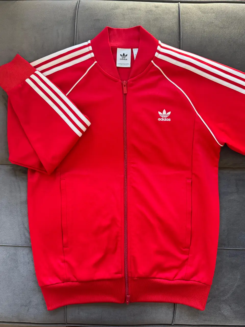 Buzo deportivo Adidas rojo con las tres tiras blancas en las mangas. Cierre frontal y logo bordado. Ideal para un look casual y deportivo.