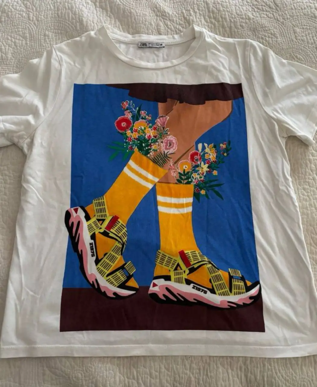 Remera blanca de algodón con estampado artístico de piernas con sandalias y flores. Ideal para un look casual y moderno.