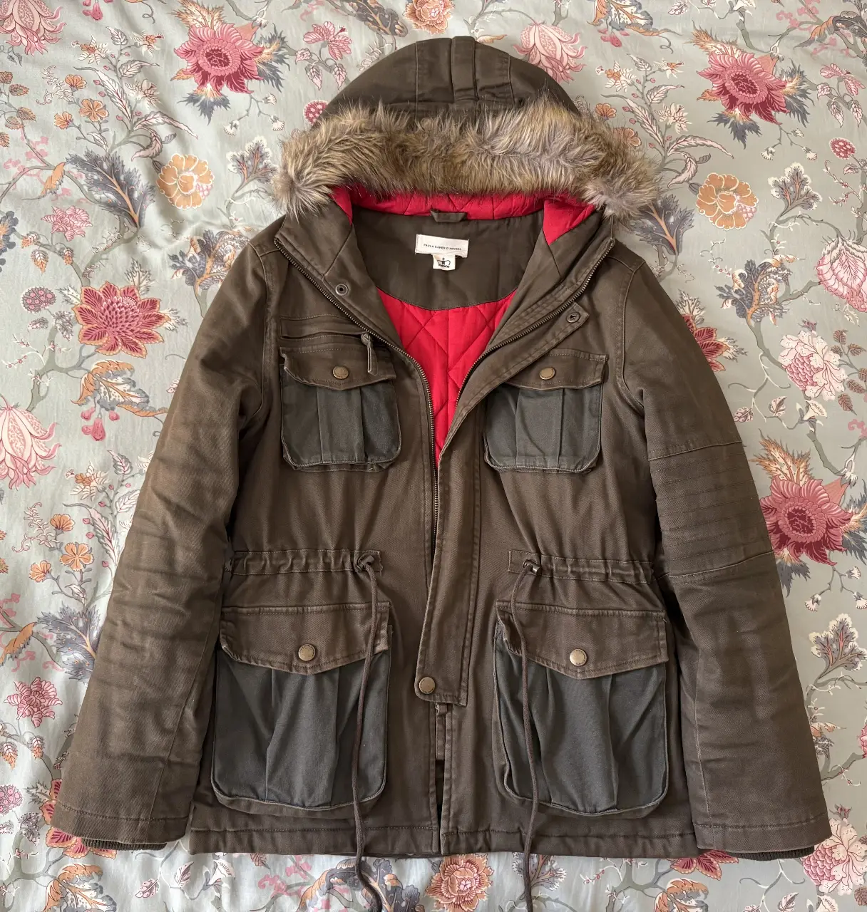 Campera Paula Cahen D'Anvers, estilo parka, color marrón oscuro con detalles en verde oscuro. Muy abrigada, no es una campera liviana. Posee capucha con borde de piel sintética, forro interior rojo acolchado y múltiples bolsillos. Ideal para el invierno. 100% algodón. 