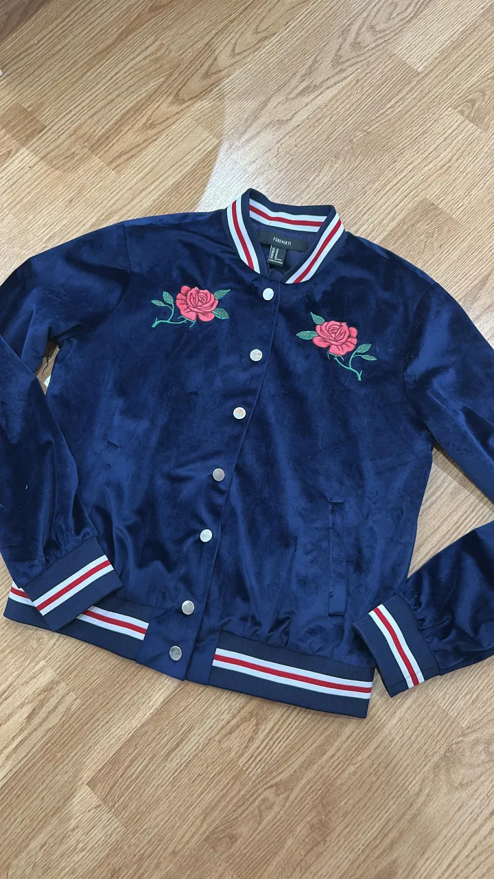 Campera bomber azul marino con bordado de rosas rojas en el frente y la inscripción "Heart Breaker" en la espalda. Posee puños y cuello a rayas rojas, blancas y azules.