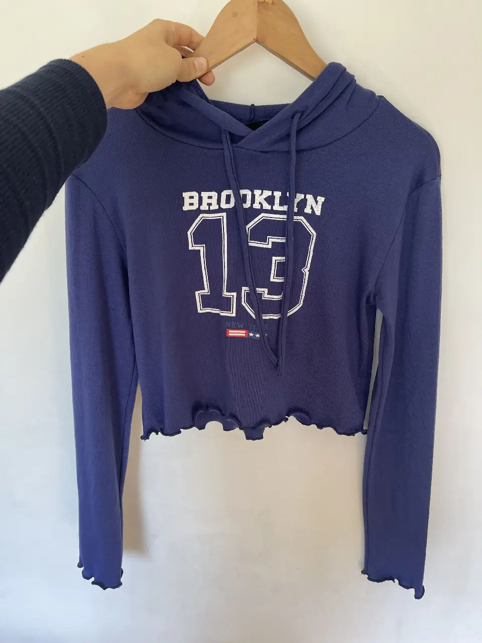 Buzo crop con capucha, color azul marino con estampado "BROOKLYN 13" en blanco. Detalles de diseño con borde ondulado en el bajo y mangas largas. Ideal para un look casual y urbano.