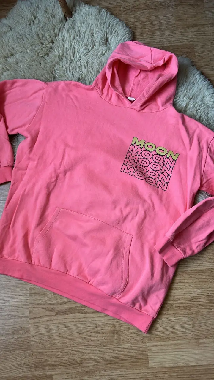 Buzo canguro rosa con capucha y bolsillo delantero. Estampado "MOON" en el frente y diseño de planeta en la espalda. Ideal para un look casual y cómodo.