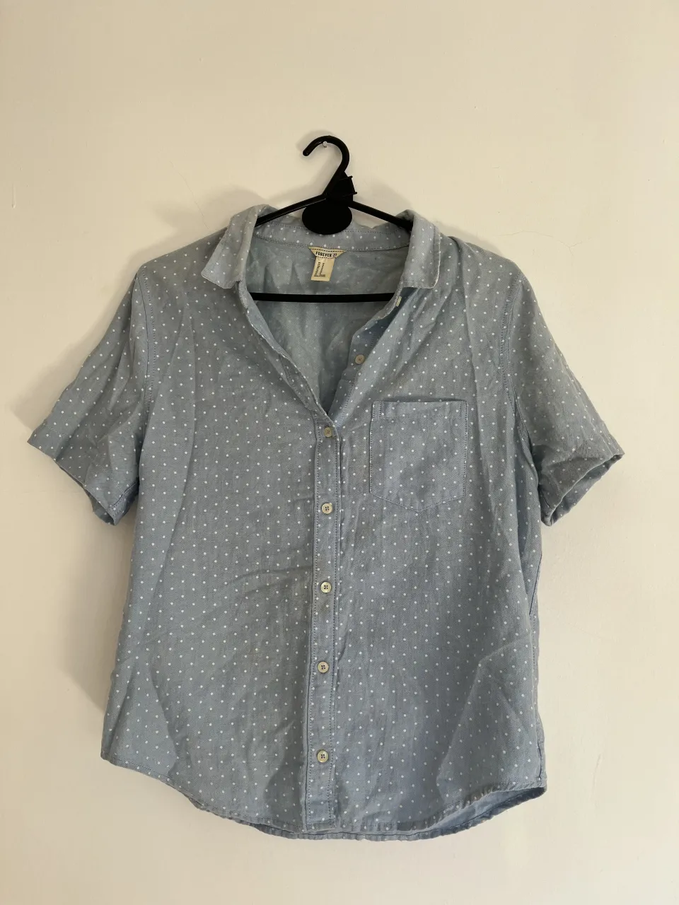 Camisa de manga corta con estampado de lunares blancos sobre fondo celeste. Posee cuello camisero, botones al frente y un bolsillo en el pecho. Ideal para un look casual y fresco.