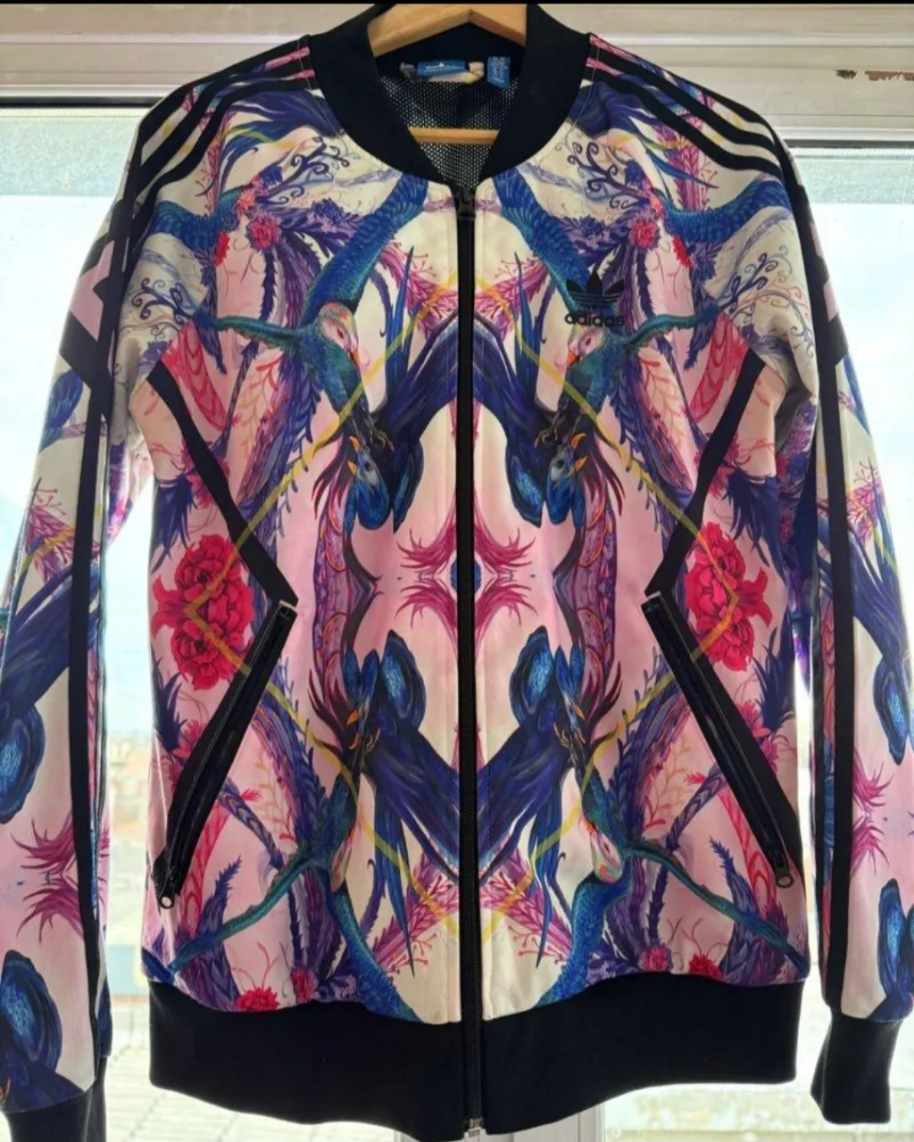 Campera deportiva estampada con diseño de aves y flores. Tiene cierre al frente, cuello redondo y puños elastizados. Las mangas tienen las tres tiras características de la marca.