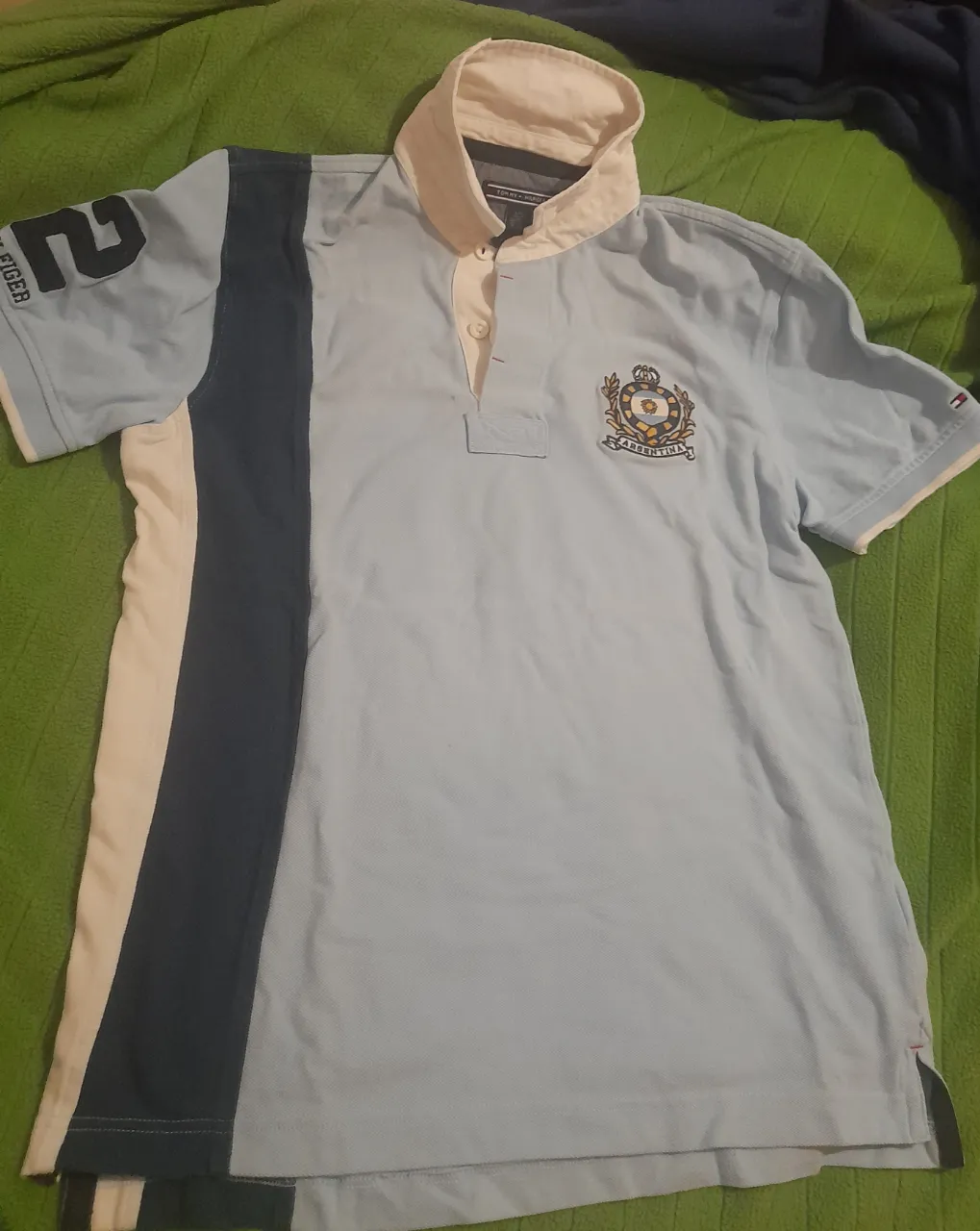 Remera de rugby Tommy Hilfiger celeste con detalles en blanco y azul marino. Cuello polo con logo bordado y número 2 en la manga. Talle L.